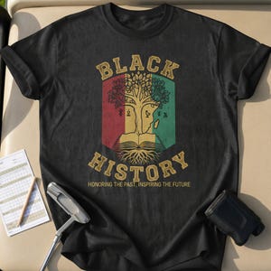 Può includere: T-shirt nero con un design grafico. Il design include le parole "BLACK HISTORY" in lettere dorate sopra un'illustrazione di un albero con un volto e un libro aperto. Il testo "HONORING THE PAST, INSPIRING THE FUTURE" è sotto.