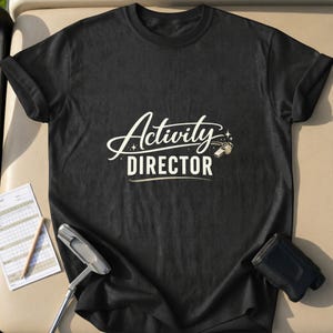 Könnte beinhalten: Schwarzes T-Shirt mit dem weißen Schriftzug "Activity Director". Ein Golfschläger, eine Scorekarte und ein Entfernungsmesser sind im Bild zu sehen. Das T-Shirt ist aus weichem Material.