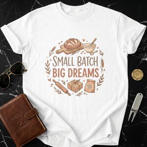 Peut inclure: T-shirt blanc avec une illustration circulaire représentant des outils et des ingrédients de boulangerie. Le motif comprend un pain, un fouet, un rouleau à pâtisserie et un sac de farine. Le texte "SMALL BATCH BIG DREAMS" est également inclus.