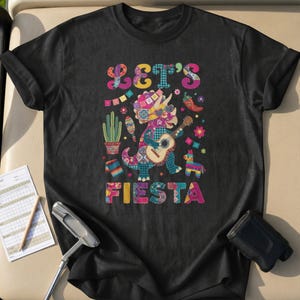 Può includere: T-shirt nero con un design colorato "Let's Fiesta". La grafica presenta un dinosauro che suona la chitarra, circondato da elementi festivi come un cactus, peperoncini e una piñata. Il design è vivace e accattivante.