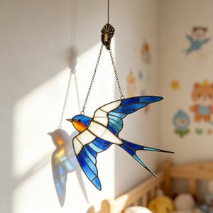 May include: A stained glass swallow bird with blue, white, and orange details hangs from a chain. The bird's wings are spread in flight, and the glass catches the light, creating a colorful shadow on the wall. The bird is suspended from a decorative metal fixture.
