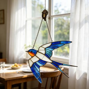 May include: A stained glass swallow suncatcher with a blue, white, and orange design. The bird is suspended by a chain in front of a window with white curtains. The swallow's wings are spread in flight.
