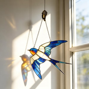 May include: Stained glass swallow bird suncatcher with blue, white, and orange hues. The bird is suspended by a chain and decorative fixture. The suncatcher is hanging near a window, casting a colorful shadow on the wall.