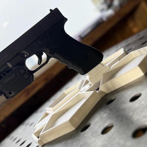 Könnte beinhalten: Eine schwarze Handfeuerwaffe mit montierter Lampe und einem cremefarbenen, geometrisch geformten Objekt. Die Handfeuerwaffe liegt auf dem Objekt, das ein maßgefertigter Halter oder eine Präsentationsvorrichtung zu sein scheint. Der Hintergrund ist eine metallische Oberfläche.