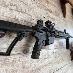 May include: Black rifle with a textured grip and adjustable stock, mounted on a wooden wall. The rifle features a scope and other tactical accessories. The background is a light-coloured, weathered wood.