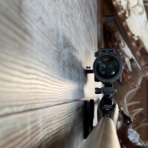 May include: A rifle with a mounted scope, positioned against a wooden wall. The scope is black with a green lens. The rifle's barrel is a light brown color. The background includes a wooden shelf and animal skulls.