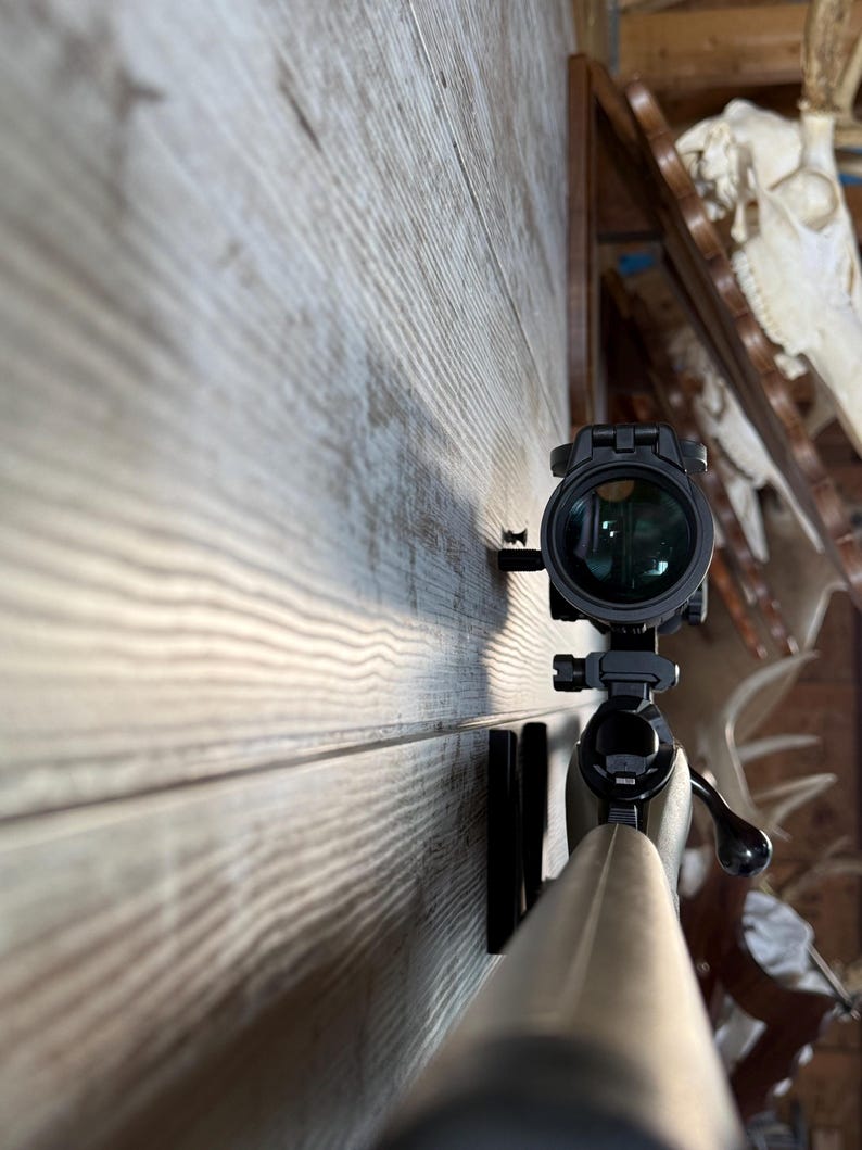 May include: A rifle with a mounted scope, positioned against a wooden wall. The scope is black with a green lens. The rifle's barrel is a light brown color. The background includes a wooden shelf and animal skulls.