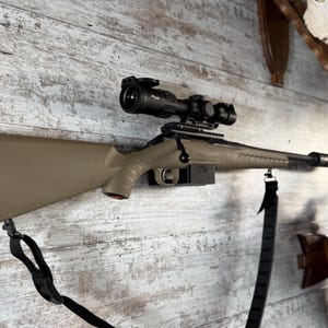 May include: A tan and black rifle with a scope mounted on a wooden wall. The rifle has a black strap and a black scope. The stock is tan, and the barrel is black. Hunting and sporting goods.