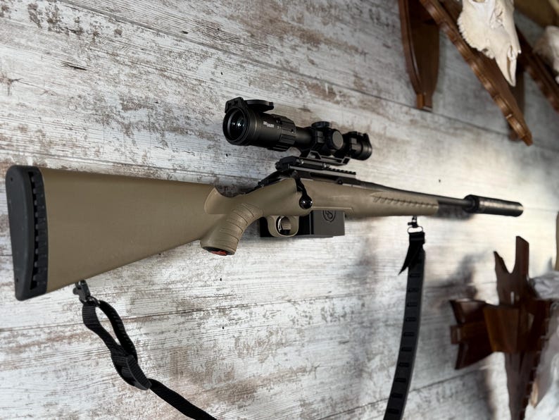 May include: A tan and black rifle with a scope mounted on a wooden wall. The rifle has a black strap and a black scope. The stock is tan, and the barrel is black. Hunting and sporting goods.