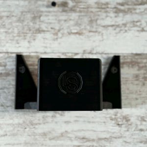 May include: A black, rectangular wall-mounted shelf with a decorative emblem. The shelf is flanked by two black, triangular brackets. The background is a light-colored, weathered wood.