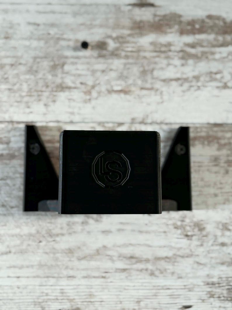 May include: A black, rectangular wall-mounted shelf with a decorative emblem. The shelf is flanked by two black, triangular brackets. The background is a light-colored, weathered wood.
