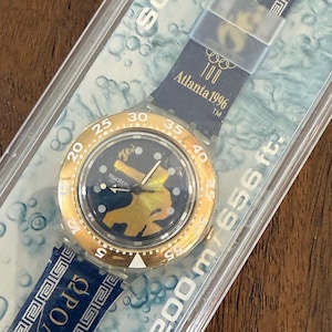 Puede incluir: Un reloj Swatch Scuba 200 en su embalaje original. El reloj presenta un bisel dorado, una esfera azul oscuro con un diseño dorado y una correa azul con patrones de llaves griegas blancas. El reloj está marcado "Atlanta 1996".