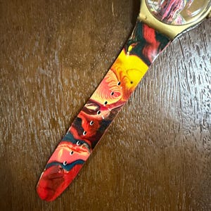 May include: A wristwatch featuring a round gold-tone case and a flexible strap decorated with a vibrant, swirling marbled pattern in shades of red, yellow, black, and blue. The watch face displays a matching abstract design under a clear crystal.
