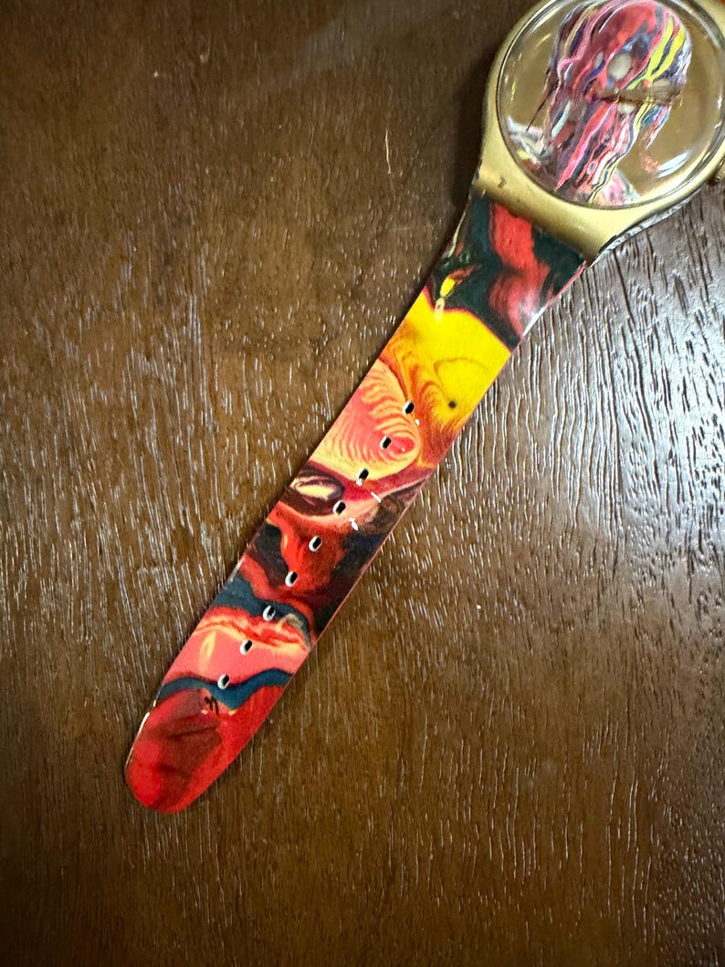 May include: A wristwatch featuring a round gold-tone case and a flexible strap decorated with a vibrant, swirling marbled pattern in shades of red, yellow, black, and blue. The watch face displays a matching abstract design under a clear crystal.