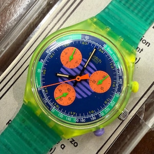 Puede incluir: Un reloj cronógrafo Swatch con caja verde neón translúcida y correa verde texturizada. La esfera azul presenta tres subesferas naranjas, un patrón de rayas moradas y manecillas verdes. El bisel incluye una escala taquimétrica con texto blanco.