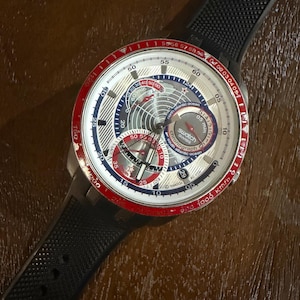 May include: A Swatch Swiss Chronograph watch with a black rubber strap. The watch face features a silver dial with red, white, and blue accents, including a map design and the text "North Atlantic Ocean". The bezel is red.