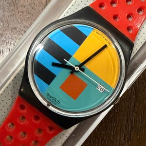 May include: A vintage Swatch wristwatch with a black case and a bright red perforated silicone strap. The watch face features a geometric design with blue, yellow, and orange blocks, black hands, and a date window displaying 28. Text reads SWATCH AG 1987.
