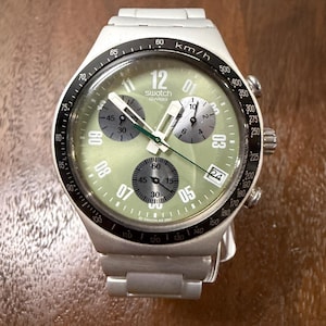 May include: A silver-toned Swatch chronograph watch with a light green dial, three dark gray sub-dials, and a black tachymeter bezel. The watch features white hour markers, a date window at the four o'clock position, and a metallic link bracelet.