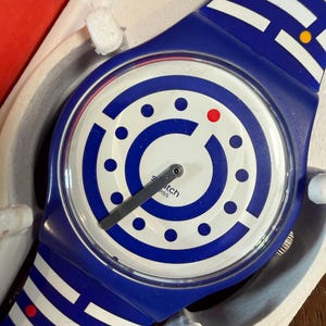 Puede incluir: Un reloj de pulsera azul y blanco con una esfera circular que presenta un patrón de laberinto de líneas y puntos azules, un punto rojo y una manecilla gris. La correa muestra un diseño geométrico de laberinto azul y blanco a juego.