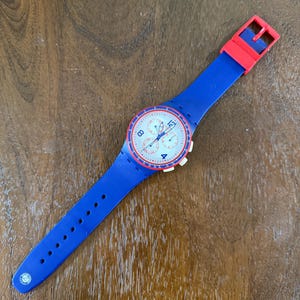 May include: A blue and red wristwatch with a white face. The watch has a blue silicone band and a red buckle. The watch face features white numbers and red accents. The watch is lying on a wooden surface.