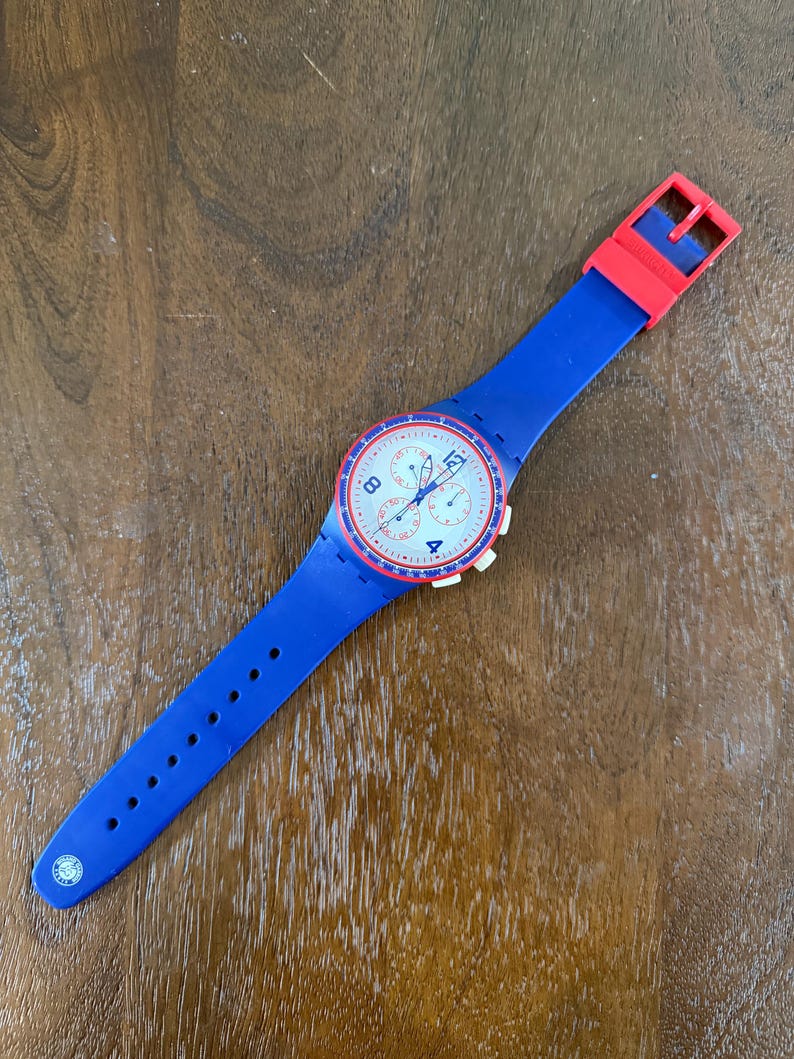 May include: A blue and red wristwatch with a white face. The watch has a blue silicone band and a red buckle. The watch face features white numbers and red accents. The watch is lying on a wooden surface.