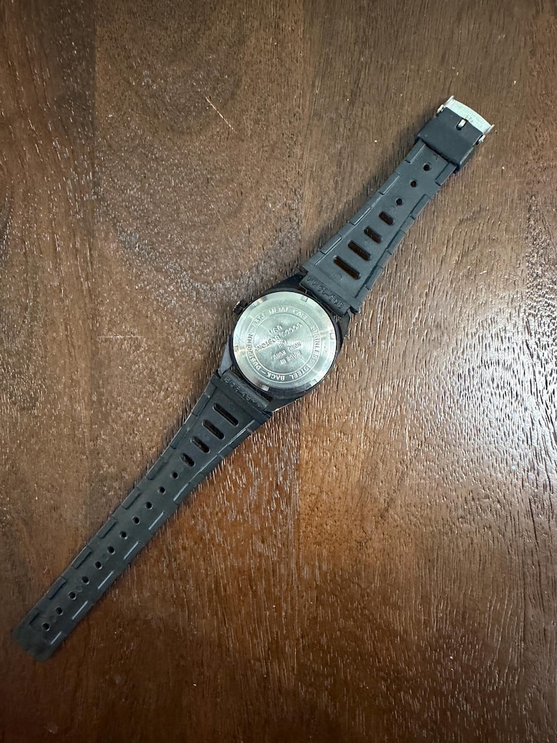 May include: A black wristwatch with a flexible rubber strap and a silver-toned stainless steel case back. The back of the watch features engraved text including the words stainless steel, water resistant, and various alphanumeric codes.