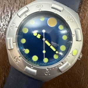 May include: A Swatch wristwatch with a dark blue dial, yellow circular hour markers, and a larger yellow circle at the twelve o'clock position. The watch features a silver-toned bezel with engraved numbers and a dark blue silicone strap.