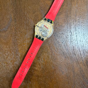 May include: A wristwatch with a transparent case back revealing the internal mechanical movement and battery. The watch features a bright red strap and a gold-toned buckle. The strap end displays a colorful, abstract pattern near the clasp.