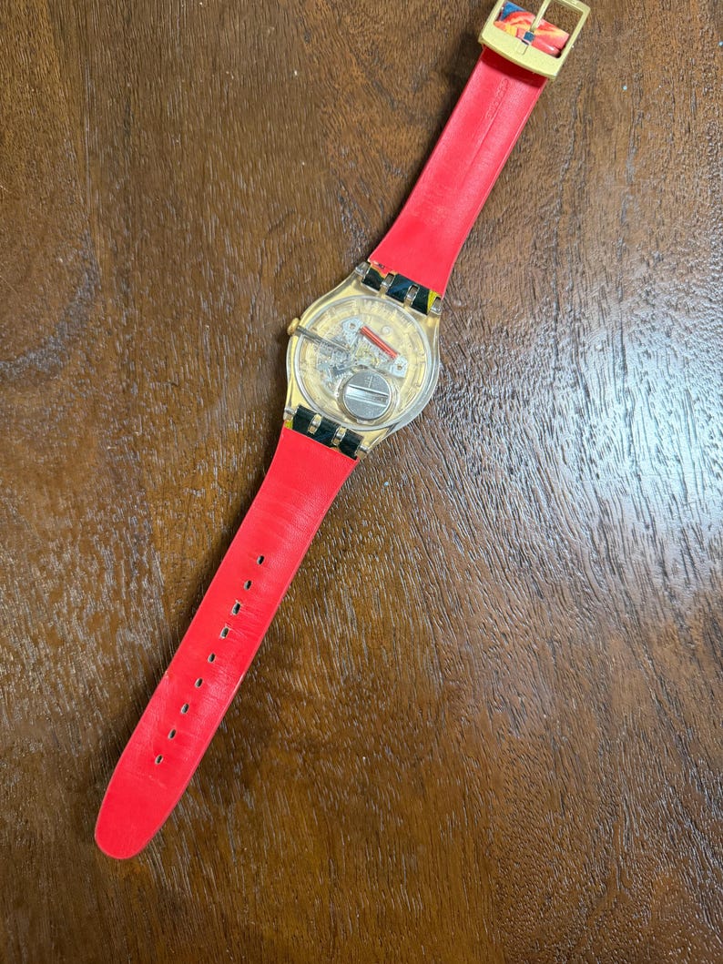 May include: A wristwatch with a transparent case back revealing the internal mechanical movement and battery. The watch features a bright red strap and a gold-toned buckle. The strap end displays a colorful, abstract pattern near the clasp.