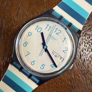May include: A Swatch wristwatch with a white face, light blue numerals, and dark blue hands. The watch features a day-date window displaying TUE 9. The strap is made of a translucent material with horizontal stripes in white, light blue, and dark blue.