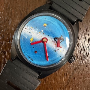May include: A black wristwatch with a blue dial featuring a space-themed graphic of a rocket, stars, and planets. The watch has bright red hour and minute hands, a black plastic strap with rectangular cutouts, and the text HONG KONG on the dial.