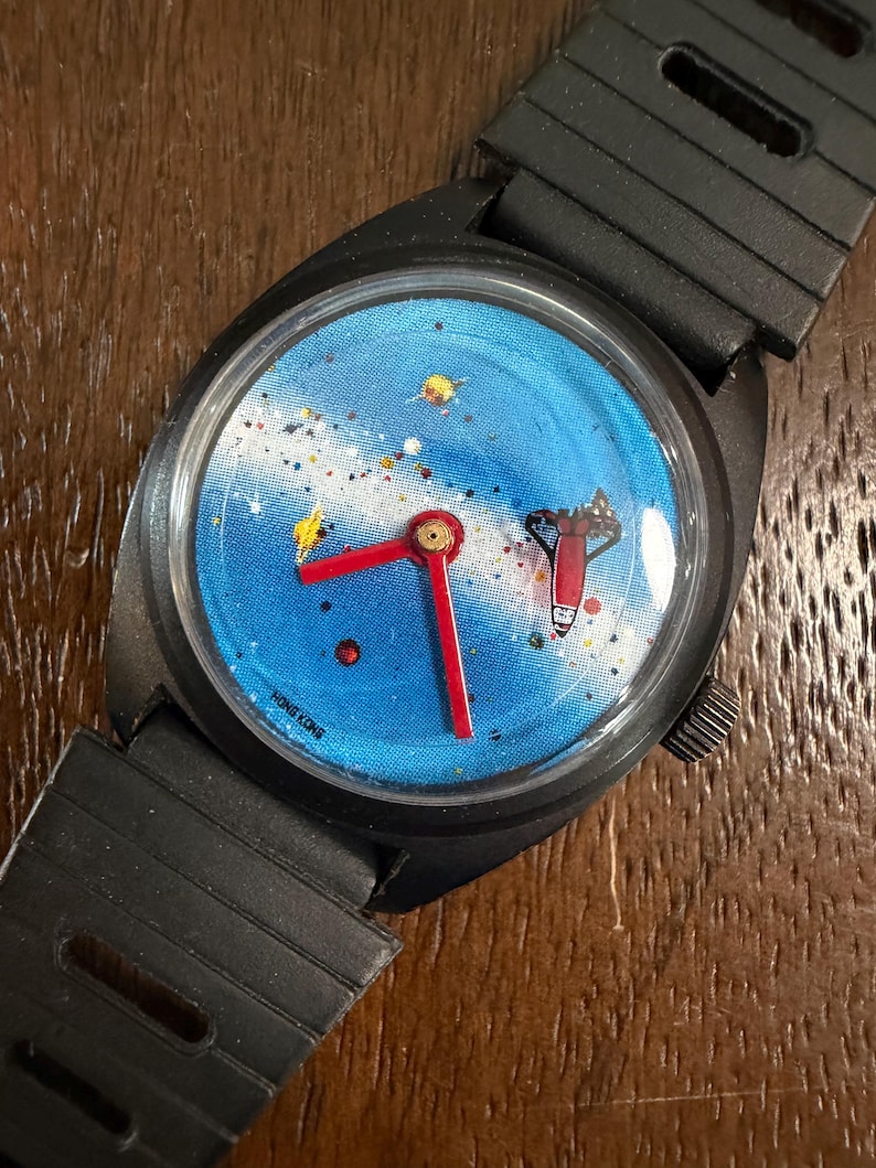 May include: A black wristwatch with a blue dial featuring a space-themed graphic of a rocket, stars, and planets. The watch has bright red hour and minute hands, a black plastic strap with rectangular cutouts, and the text HONG KONG on the dial.