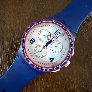 May include: A blue Swatch chronograph watch with a white face, red accents, and a blue strap. The watch face features three sub-dials and large, easy-to-read numerals. The watch has a sporty design.