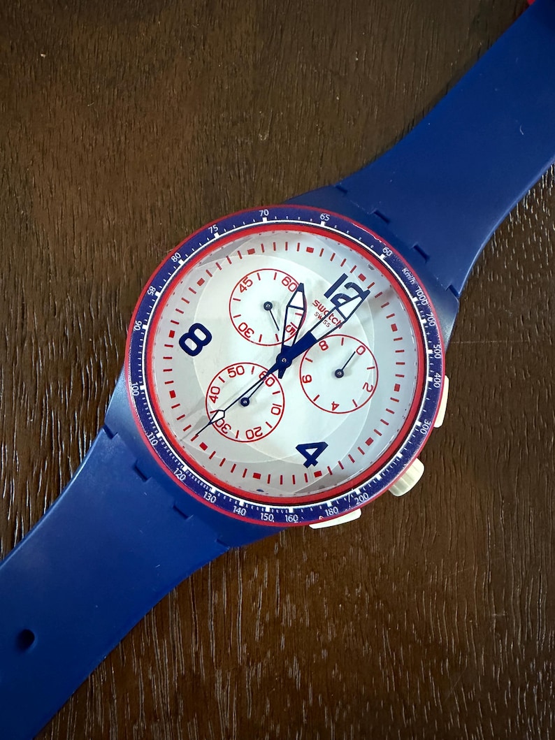 May include: A blue Swatch chronograph watch with a white face, red accents, and a blue strap. The watch face features three sub-dials and large, easy-to-read numerals. The watch has a sporty design.