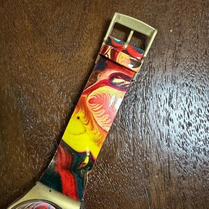 May include: A wristwatch featuring a vibrant, multi-colored marbled pattern in shades of red, yellow, and dark blue on the strap and dial. The watch includes a gold-toned metal buckle and a matching gold-toned casing around the watch face.