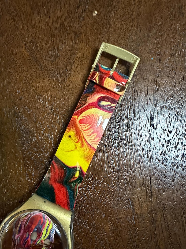 May include: A wristwatch featuring a vibrant, multi-colored marbled pattern in shades of red, yellow, and dark blue on the strap and dial. The watch includes a gold-toned metal buckle and a matching gold-toned casing around the watch face.