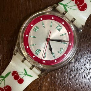 Peut inclure: Une montre-bracelet Swatch avec un boîtier en plastique transparent et un bracelet en silicone blanc orné d'un motif de cerises rouges. Le cadran présente un centre argenté, un anneau rouge à pois blancs et des chiffres verts à 3, 6, 9 et 12h.