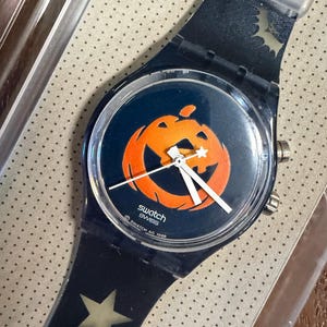 Puede incluir: Un reloj de pulsera Swatch con correa y esfera azul oscuro. La esfera presenta un gráfico de calabaza naranja. La correa muestra una estrella clara y una silueta de murciélago. El texto "swatch SWISS" y "© SWATCH AG 1998" está impreso en la esfera.