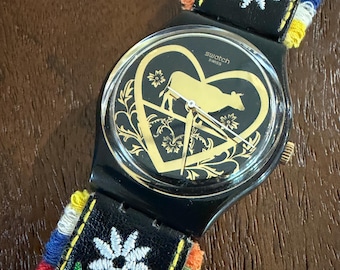 Swatch Die Glocke GB285 Originale Gentle 34 mm | Schweizer Pastorales Thema | Goldfarbenes Ziffernblatt, Armband mit Blumen bestickt, Miniatur Kuhglocke Anhänger