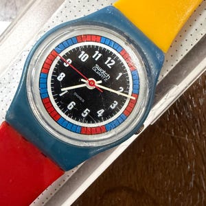 Puede incluir: Un reloj de pulsera Swatch Quartz con caja de plástico azul, esfera negra con números blancos y un anillo exterior con patrón rojo y azul. El reloj tiene una correa superior amarilla y una correa inferior roja, con manecillas blancas y segundero rojo.