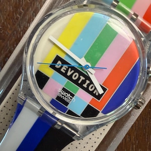 Könnte beinhalten: Eine Swatch-Armbanduhr mit einem transparenten Kunststoffgehäuse und einem Zifferblatt mit einem bunten vertikalen Streifenmuster, das an ein Fernseh-Testbild erinnert. Das Wort DEVOTION ist in Schwarz auf einem weißen Rechteck gedruckt.