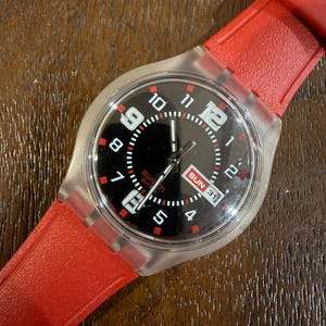 May include: A Swatch wristwatch featuring a black dial with white Arabic numerals, red accents, and a day-date window displaying SUN 31. The watch has a clear, transparent plastic case and a bright red silicone strap with a textured finish.
