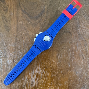 May include: A blue Swatch watch with a red buckle and strap. The watch has a white dial and a blue strap with a grid pattern. The watch is lying on a wooden surface.
