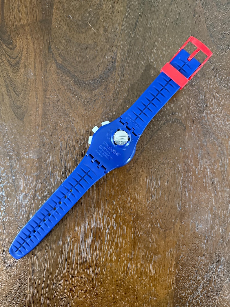 May include: A blue Swatch watch with a red buckle and strap. The watch has a white dial and a blue strap with a grid pattern. The watch is lying on a wooden surface.