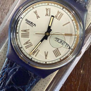 Puede incluir: Un reloj de pulsera Swatch con esfera blanca, números romanos dorados y manecillas negras. El reloj cuenta con una ventana de día y fecha que muestra SUN 11, una caja de plástico azul oscuro y una correa de cuero azul oscuro texturizada.