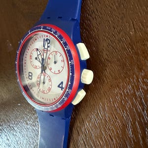 May include: A blue Swatch chronograph watch with a white face, red bezel, and blue strap. The watch features white buttons and a speedometer scale. The face displays the Swatch logo and the numbers 4 and 8.