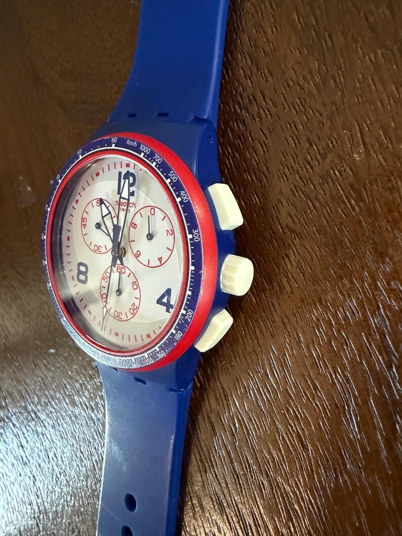 May include: A blue Swatch chronograph watch with a white face, red bezel, and blue strap. The watch features white buttons and a speedometer scale. The face displays the Swatch logo and the numbers 4 and 8.