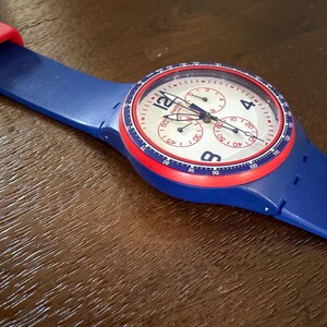 May include: A blue wristwatch with a red bezel and strap accents. The watch face is white with black numerals and three sub-dials. The watch has a blue strap and a red buckle. The watch is a chronograph style.