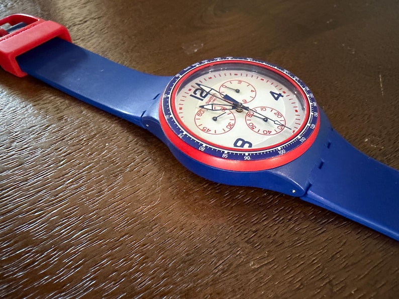May include: A blue wristwatch with a red bezel and strap accents. The watch face is white with black numerals and three sub-dials. The watch has a blue strap and a red buckle. The watch is a chronograph style.