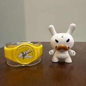 May include: A yellow wristwatch with a cartoon bear design on the face, next to a white figurine of a rabbit with a mustache. The watch has a yellow band and a clear plastic display stand. The rabbit figurine has black eyes and a brown mustache.